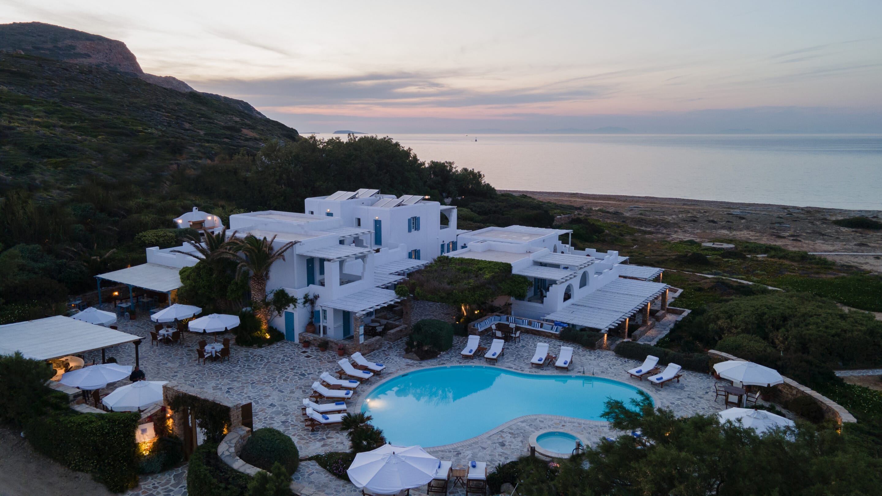 Aerial view of Villa Marandi Naxos Luxury Suites, nestled amidst lush greenery with a backdrop of the crystal-clear turquoise waters of Naxos. The hotel's pristine white buildings stand out against the vibrant Mediterranean landscape, inviting guests to indulge in a luxurious retreat.